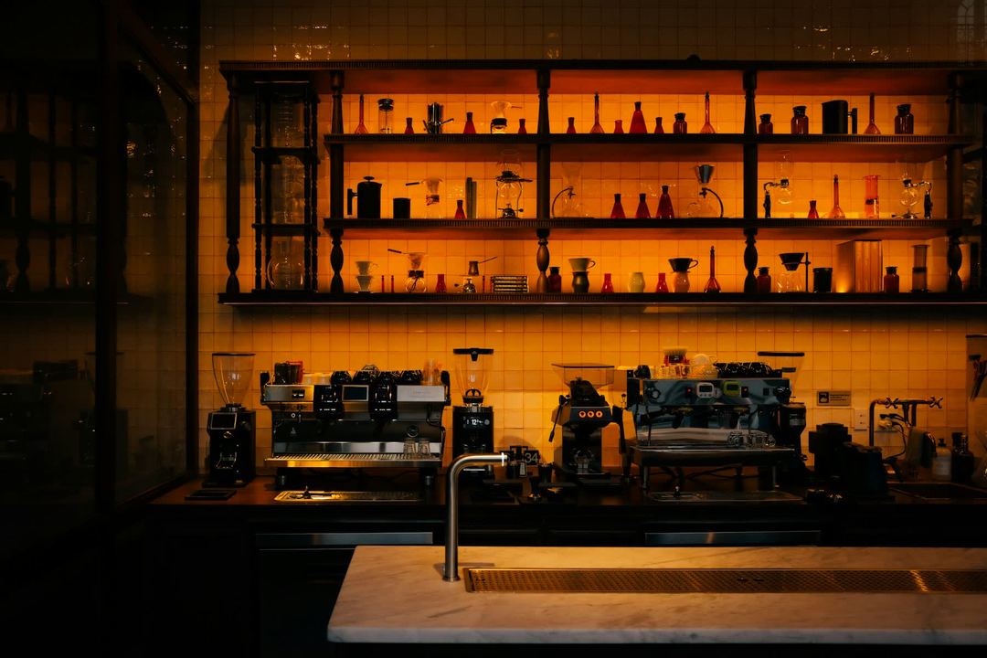A coffee bar after hours: glass brewers on lit amber shelves over two espresso machines and their grinders, a marble counter in front.
