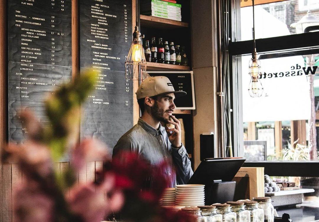 A shop owner at the register, daylight from the window behind him, the menu board on the wall.